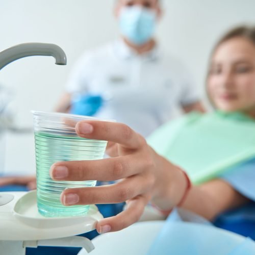 Female patient in dental chair reaching for disposable cup with mouthwash in presence of doctor